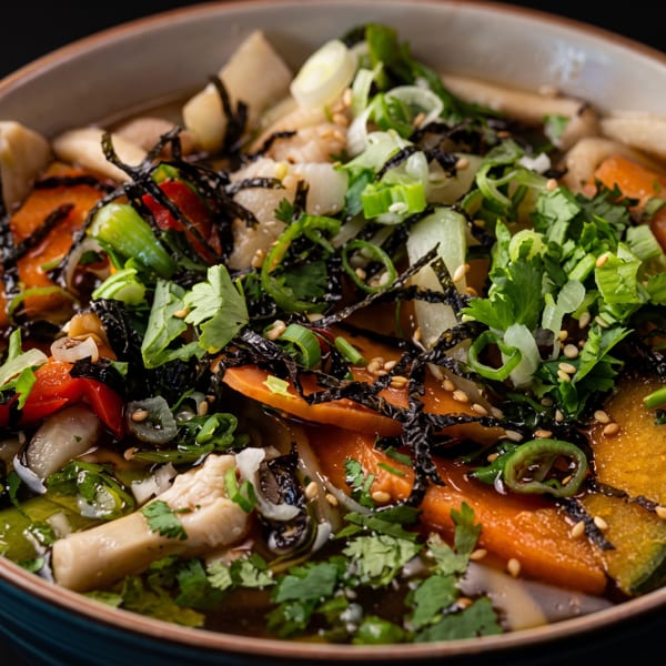 Close-up of a vibrant Asian noodle soup with vegetables, mushrooms, nori, and sesame seeds.